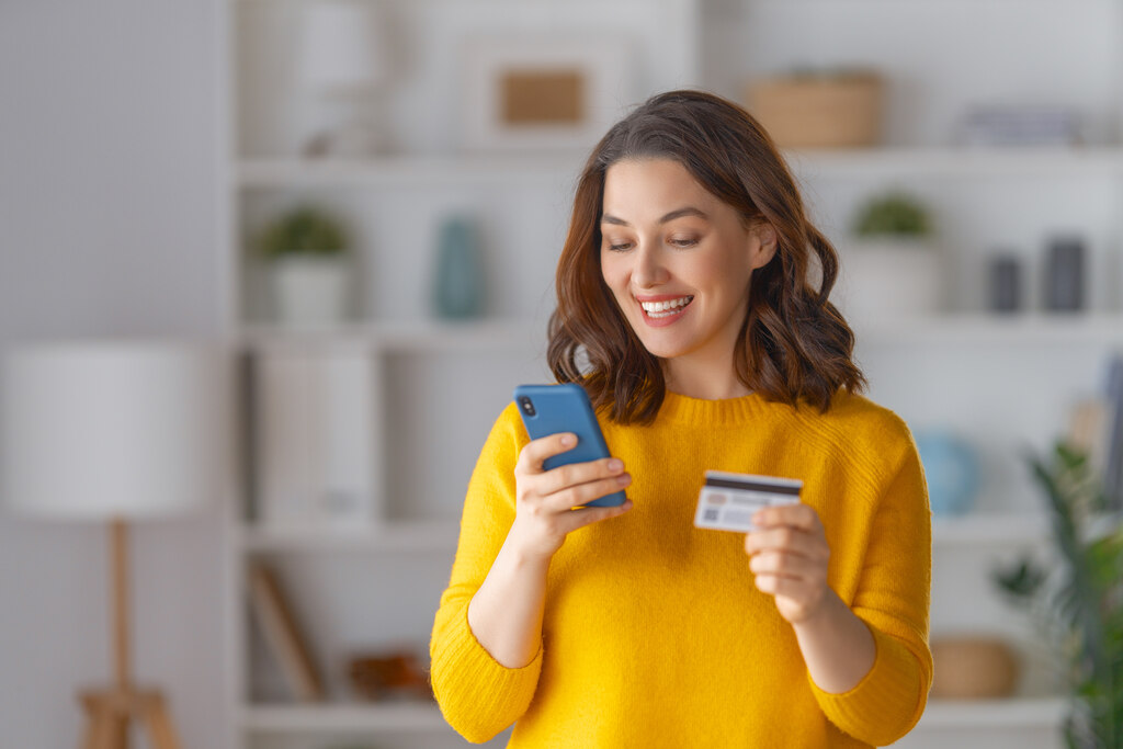 mulher sorrindo em pé segurando cartão de crédito e celular